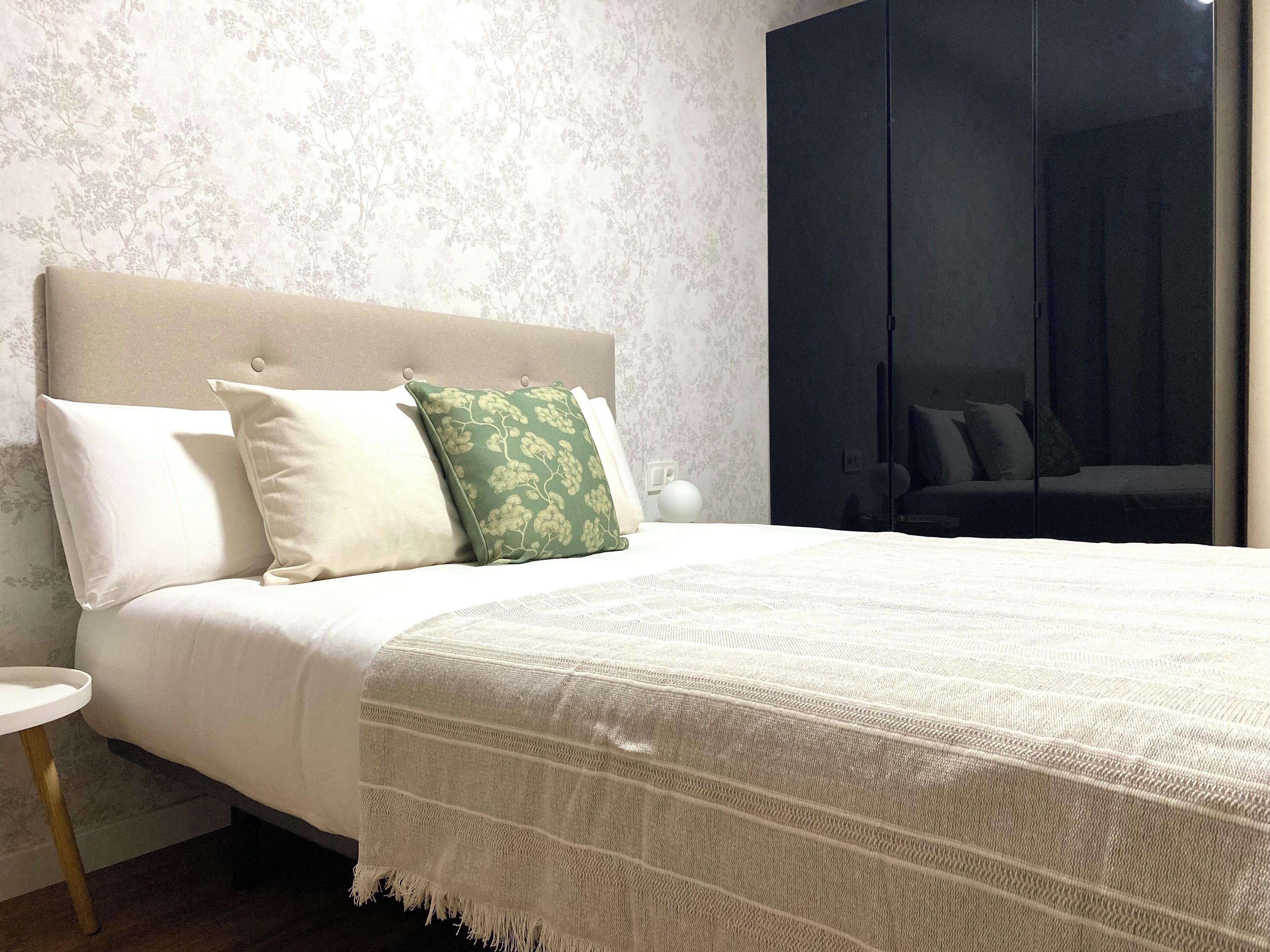 Una cama bien hecha con un cojín blanco y verde estampado, una manta blanca y una mesita de noche de madera con una lámpara blanca a un lado.