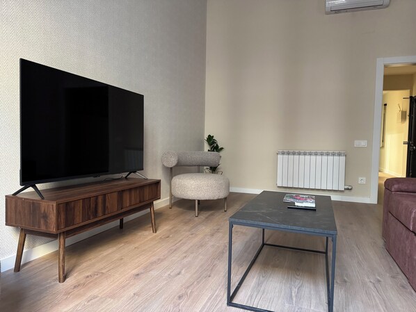 A living room with a flat-screen TV on a wooden stand, a grey upholstered chair, a small coffee table, and a radiator.