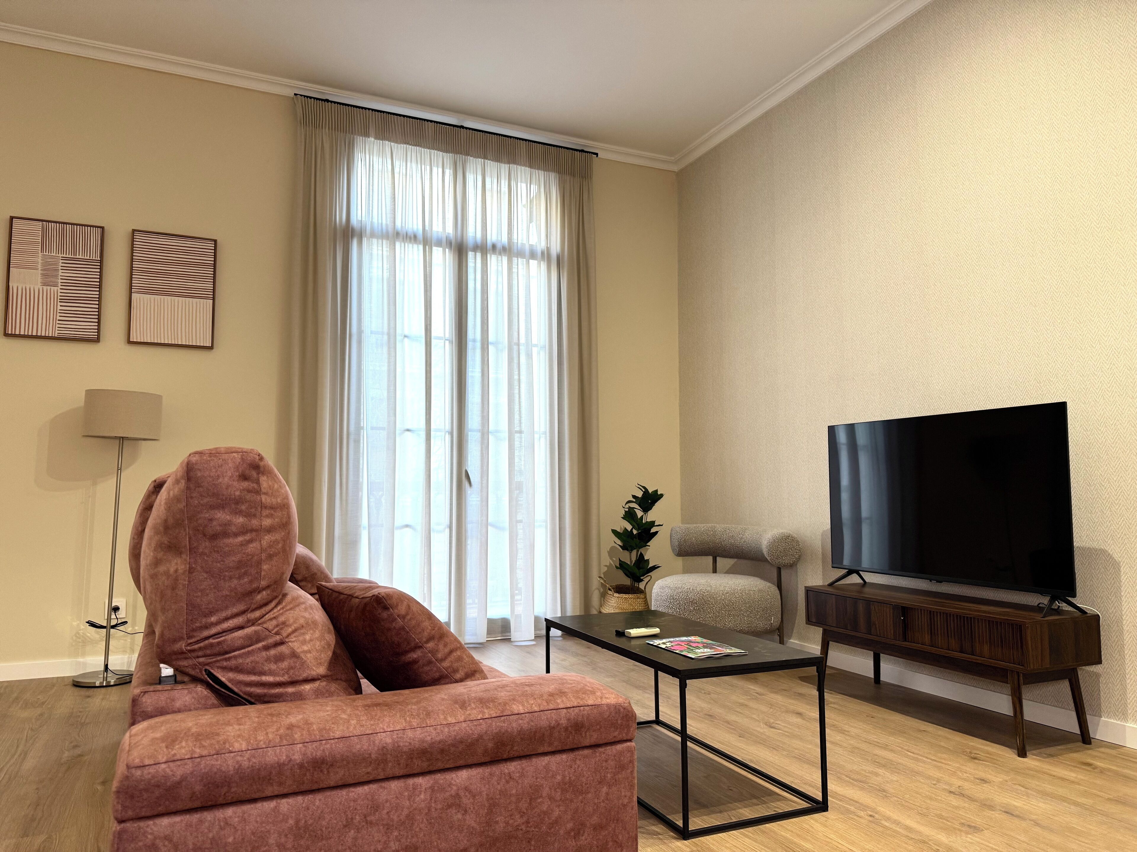 Een moderne woonkamer met een bank, een salontafel, een televisie op een houten plank, een vloerlamp en een groot raam met gordijnen.