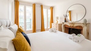Quarto de hotel com uma cama grande, lareira, espelho redondo e vista da cidade através de janelas amplas com cortinas amarelas.