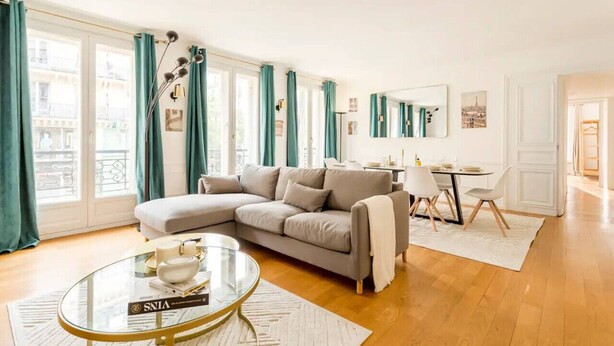 Uma sala de estar com sofá bege, uma mesa de centro de vidro e um espaço de jantar com uma mesa pequena e cadeiras.