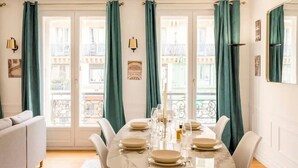 Uma área de refeições com uma mesa de mármore posta para quatro pessoas, cortinas verdes e vista para varanda.