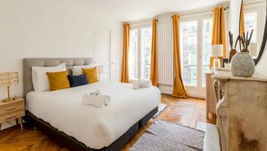 Quarto de hotel com uma cama grande, mesa de cabeceira, cômoda, espelho e janela com cortinas.