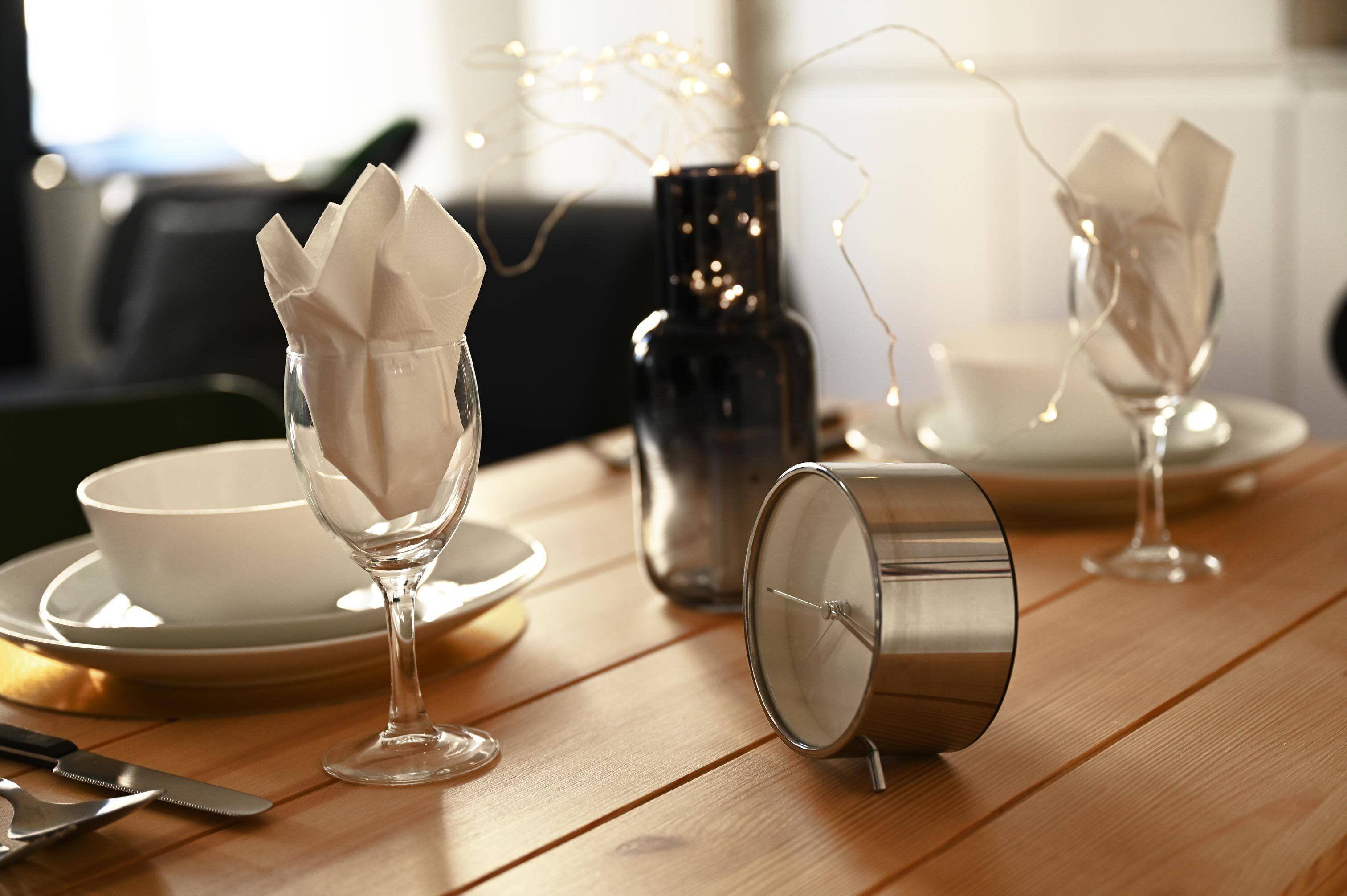 Une table à manger dressée avec de l’assiette blanche, de la verrerie transparente et un récipient en métal.