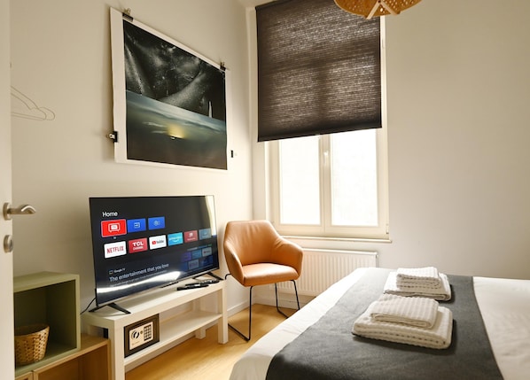 A modern hotel room with a large flat-screen TV displaying various streaming services, a comfortable chair, a neatly made bed with towels, and a large window with a view of the night sky.
