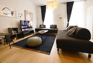 A modern living room with a sofa, ottoman, and coffee table. There is a TV on a stand, a lamp, and artwork on the wall.