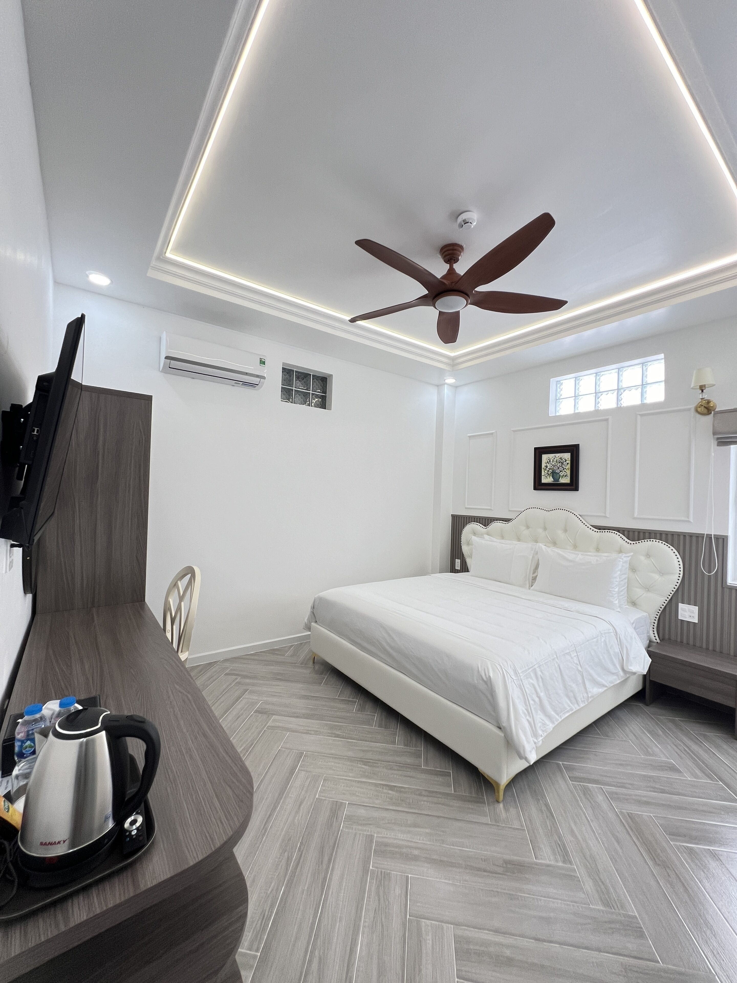 Un dormitorio moderno con cama, ventilador de techo, televisor montado en la pared y un escritorio de madera con hervidor.