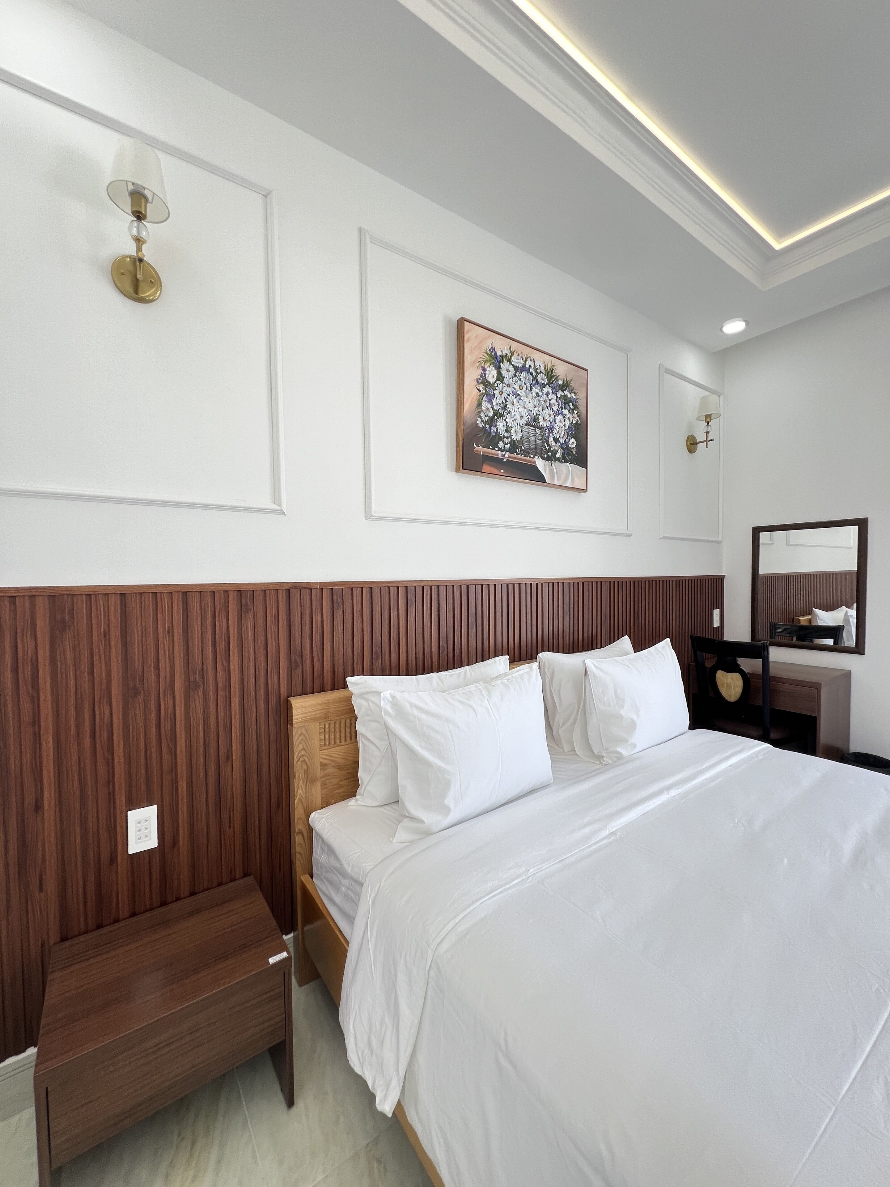 A neatly arranged hotel room with a wooden headboard, a bedside table, a bench, and a large bed with white bedding.