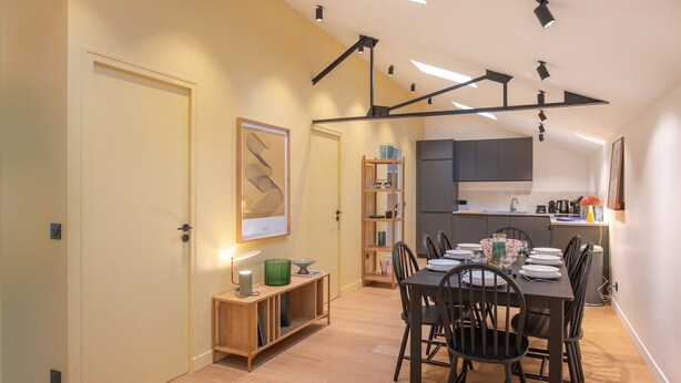 A modern kitchen with dark cabinets, a dining area with a wooden table and chairs, and a small wooden side table with a lamp.