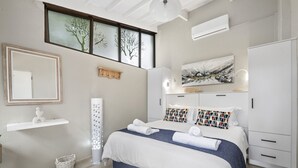 A modern bedroom with a large bed, a bedside table, a wardrobe, a window with frosted glass, and a wall-mounted air conditioner. - San Lameer Villa 2412 (Southbroom)
