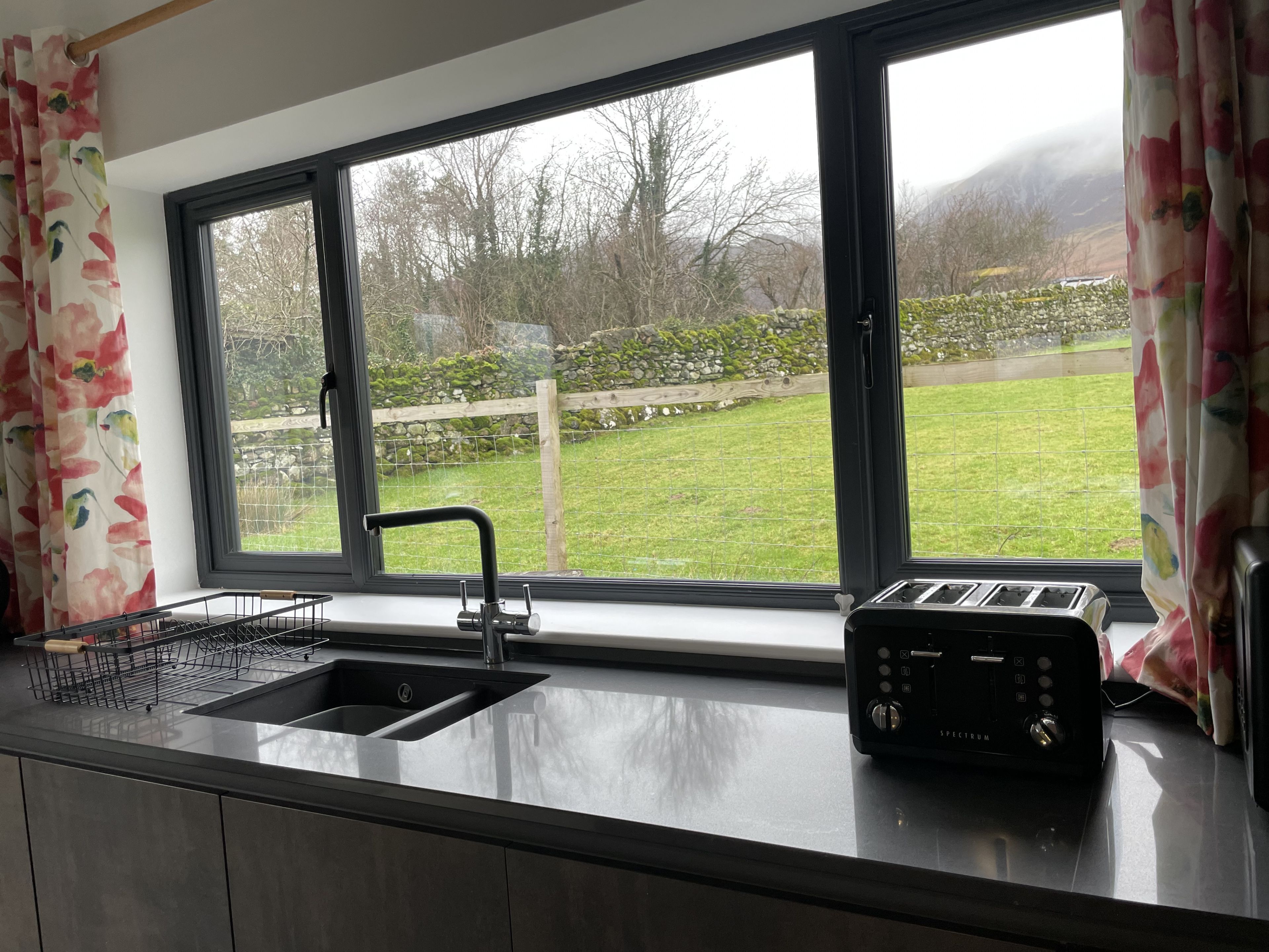A kitchen with a large window, a sink, a toaster, and a wooden fence visible outside.
