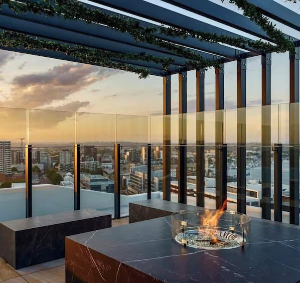 A modern outdoor seating area with a fire feature, glass railings, and a city view.