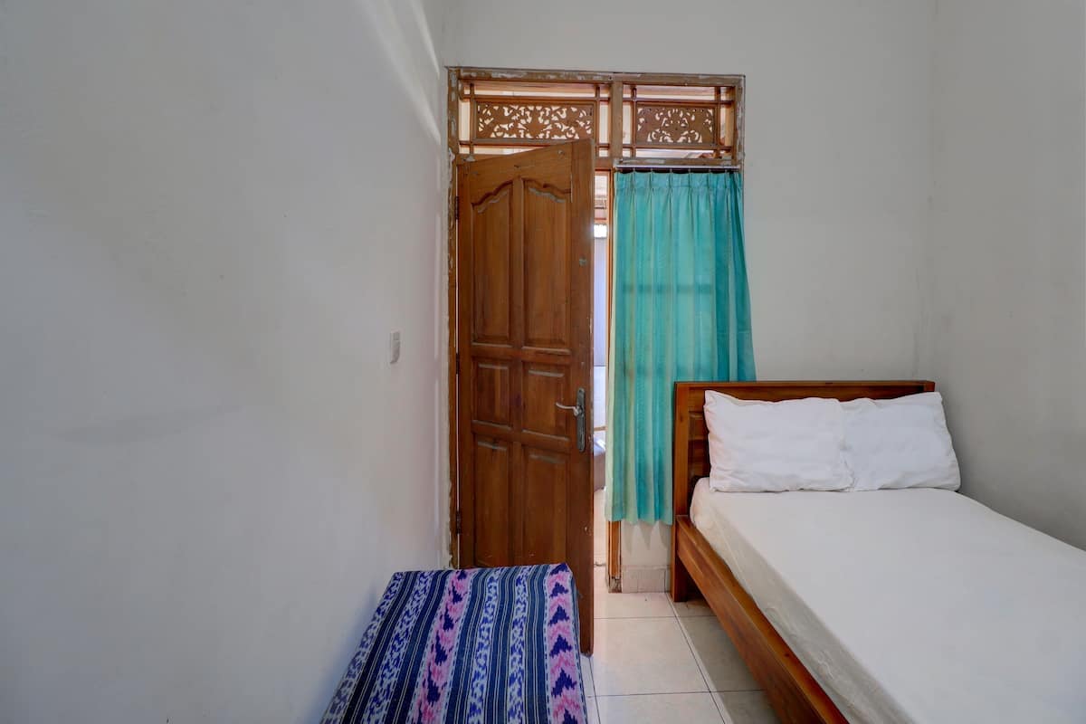 A small, narrow room with a wooden door, a bed with white bedding, and a patterned bench.