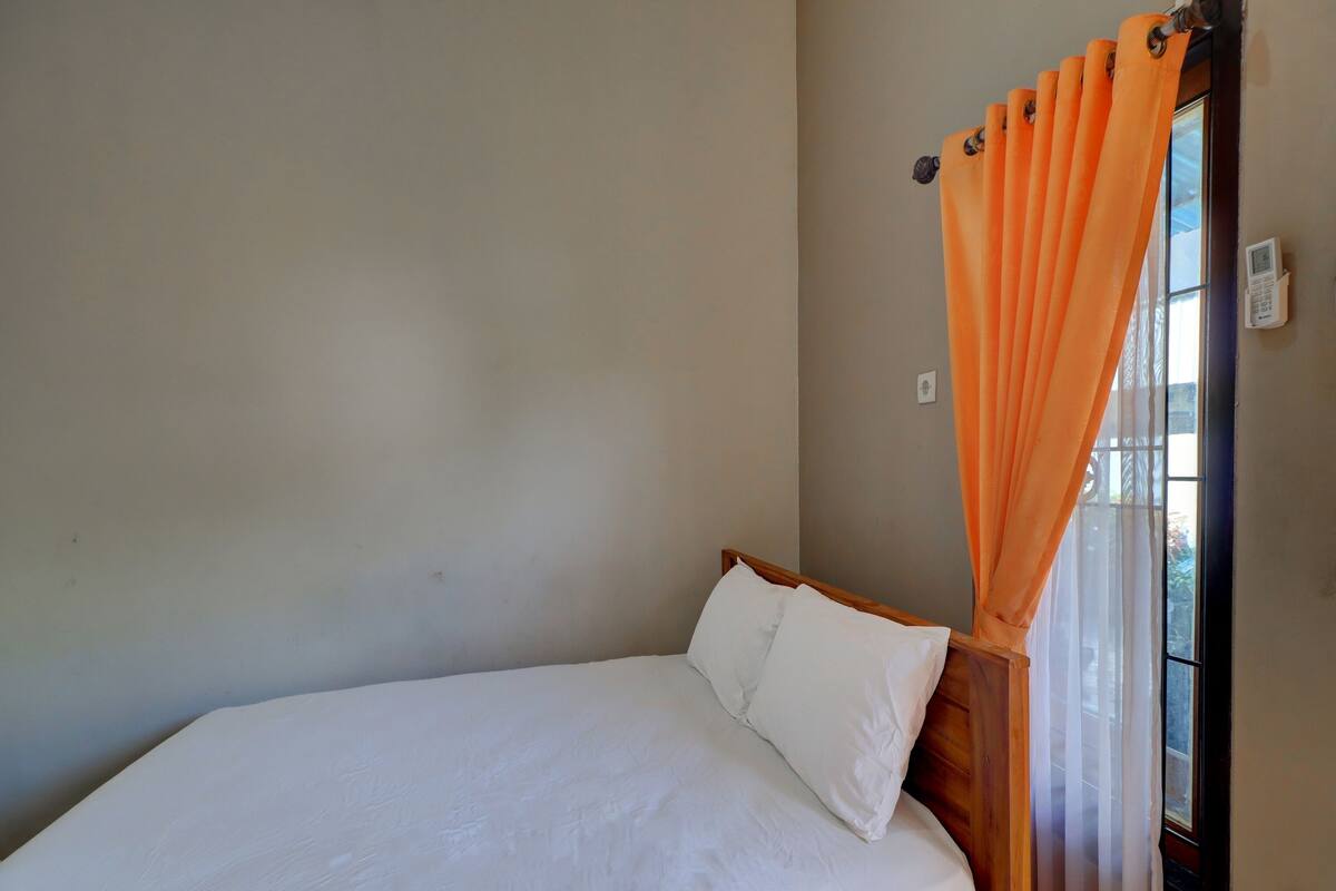 A single bed with white bedding in a room with plain walls and a window with orange curtains.