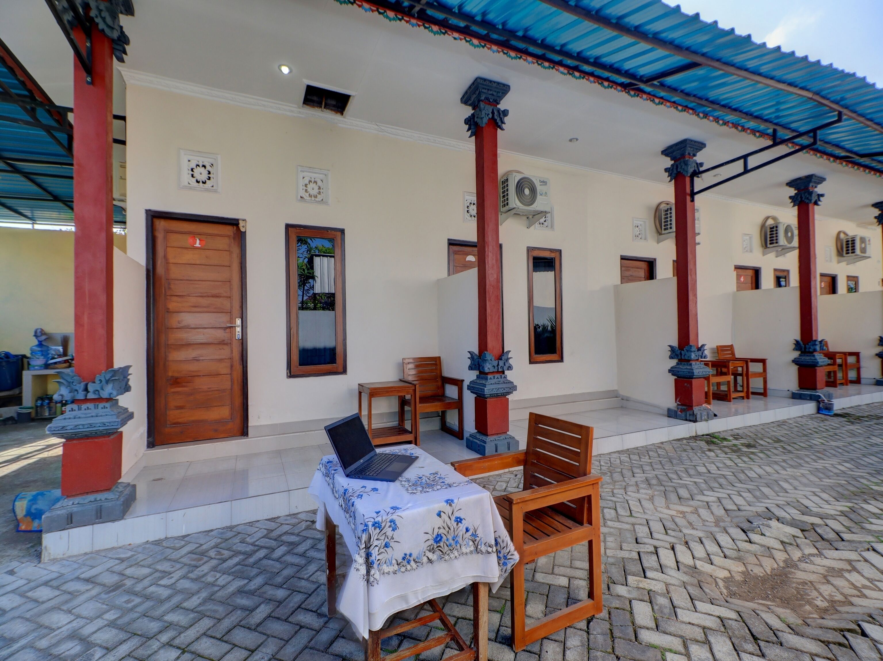 A covered outdoor seating area with wooden chairs and a table, a wooden door, and a wall-mounted air conditioning unit.