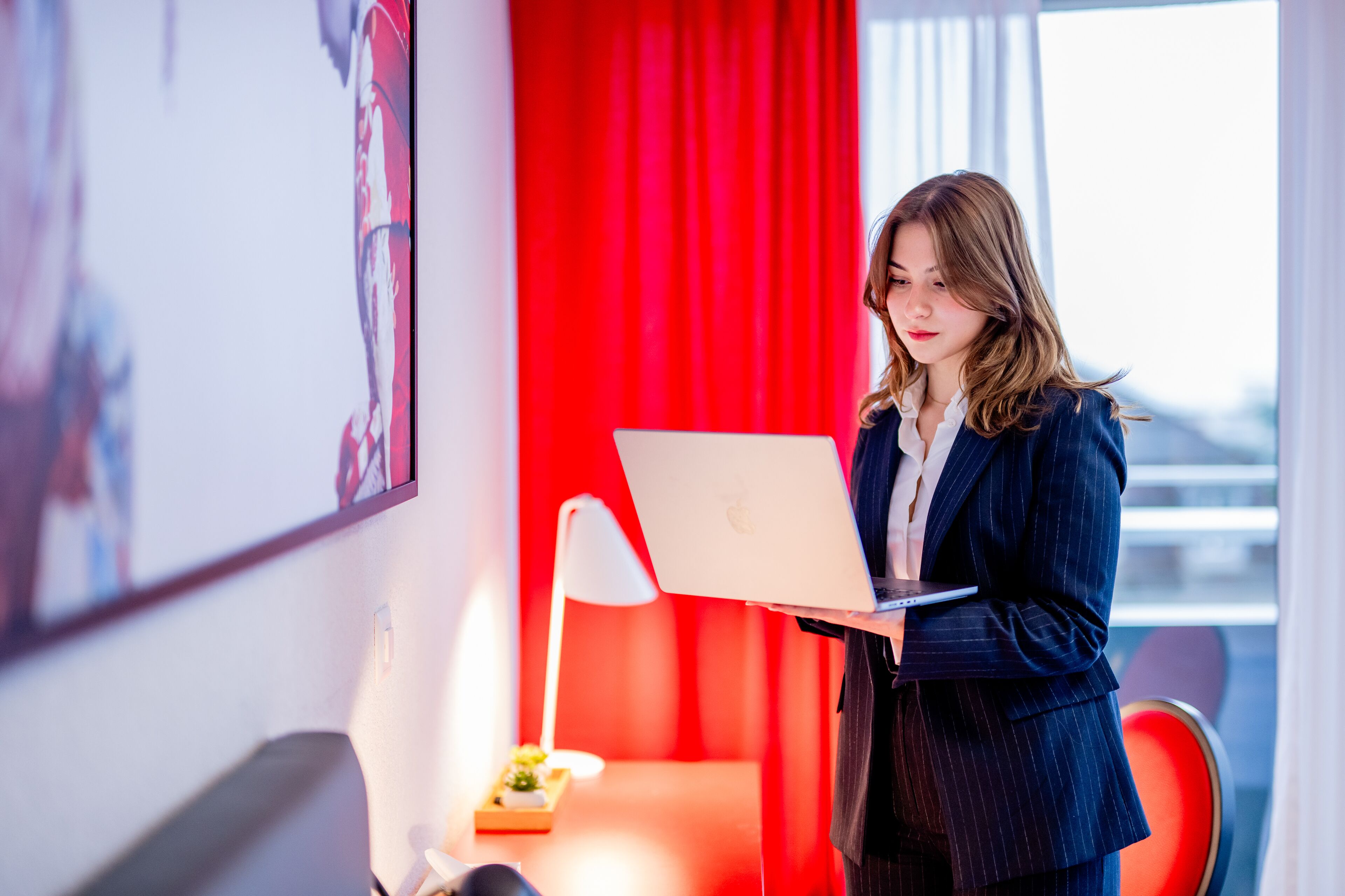 A person in a business suit holding a laptop in a room with a red curtain, a desk lamp, and a small plant.