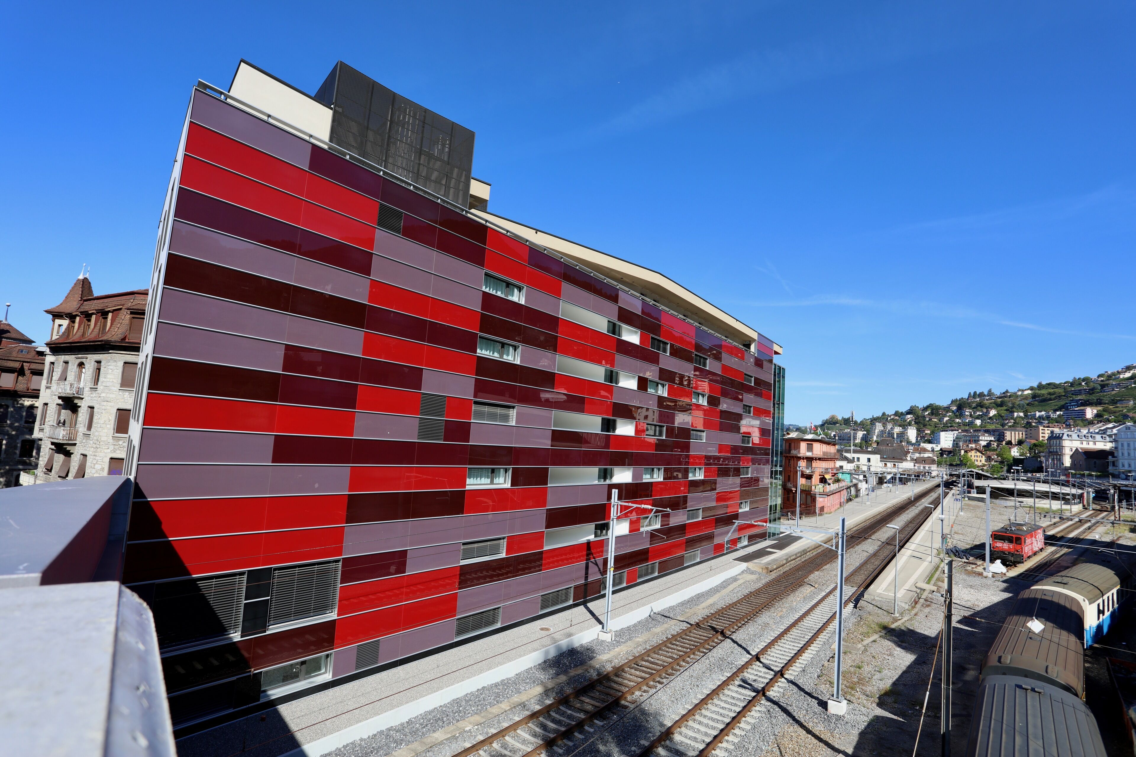 Ein modernes Gebäude mit roter und grauer Fassade, in der Nähe von Gleisen, mit einem Zug im Hintergrund.