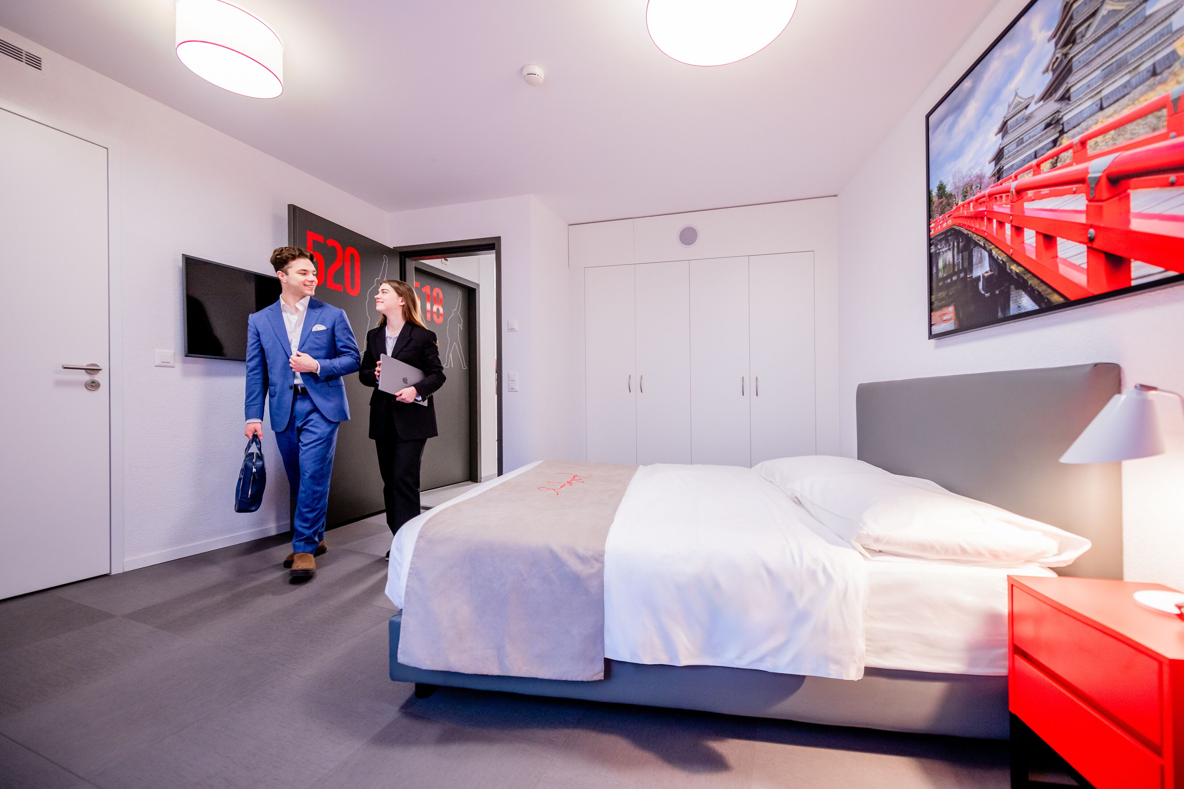 Une chambre d’hôtel moderne, équipée d’un lit, d’une table de chevet rouge, d’une télévision et d’une porte portant un numéro.