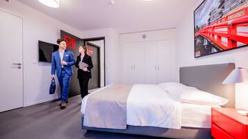 A modern hotel room with a bed, a red nightstand, a TV, and a door with a number sign.