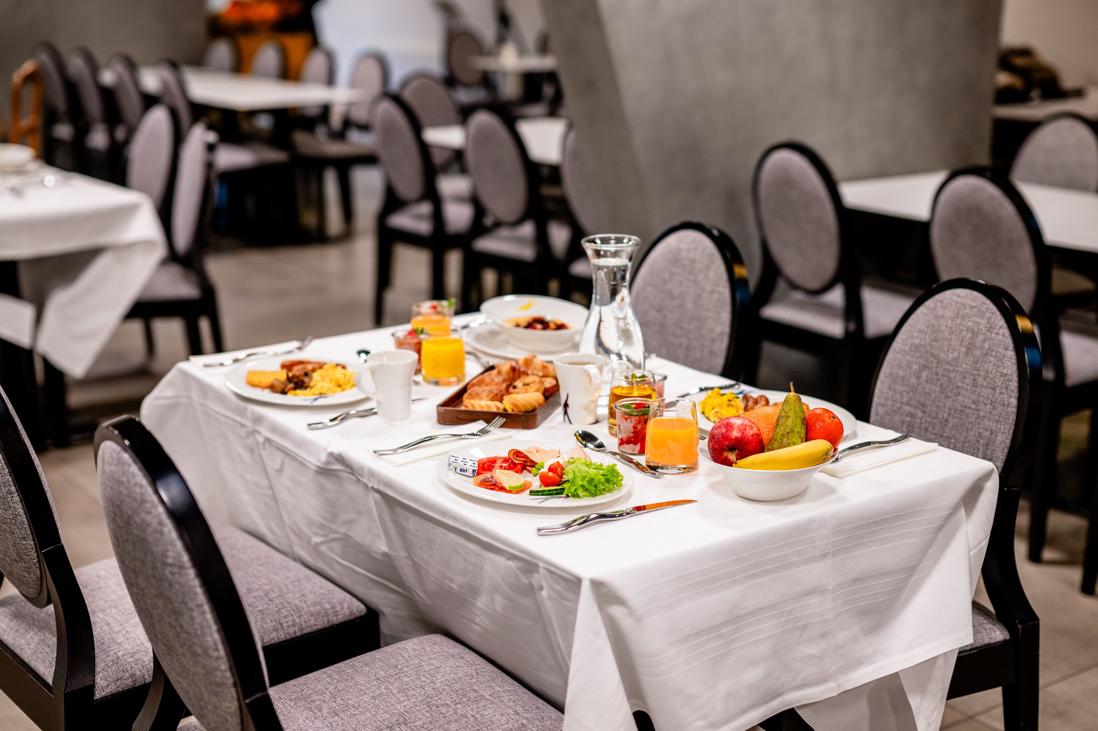 A dining table set with breakfast items, including fruits, vegetables, and beverages, in a hotel dining area with multiple tables and chairs.