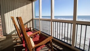 A covered outdoor seating area with wooden chairs and a red cushion, overlooking a beach with a clear view of the ocean. - Ocean's Lullaby Oceanfront Dunes Room 2nd Floor (Pawleys Island)