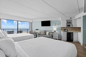 A hotel room with two beds, a kitchenette, a flat-screen TV, and a balcony with a view of the sea.