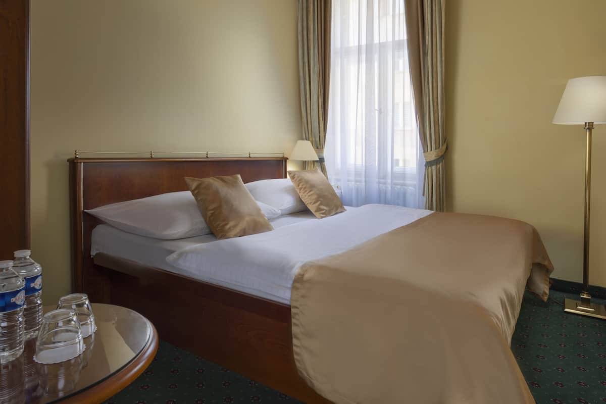 A hotel room with a bed, a bedside table with water bottles and glasses, a lamp, and a view of the outdoors through curtains.