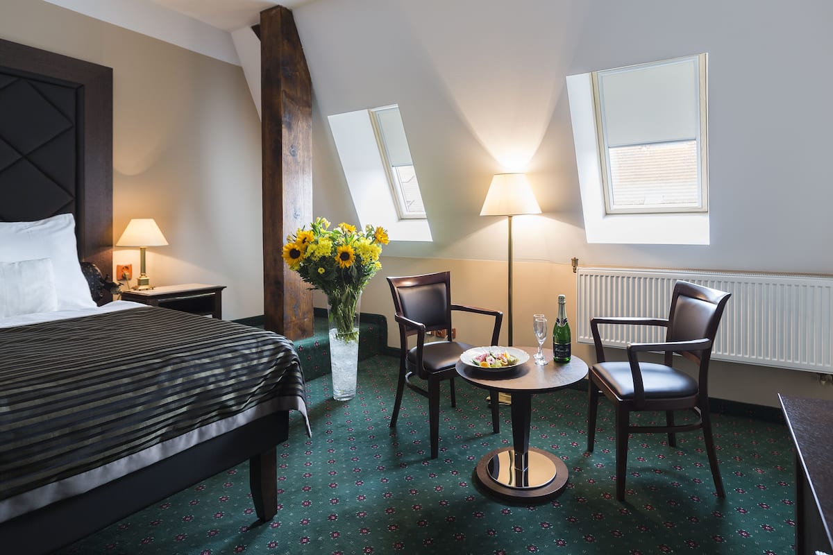 A hotel room with a bed, two chairs, a small table with a bottle and glass, a vase with flowers, and two windows with blinds.