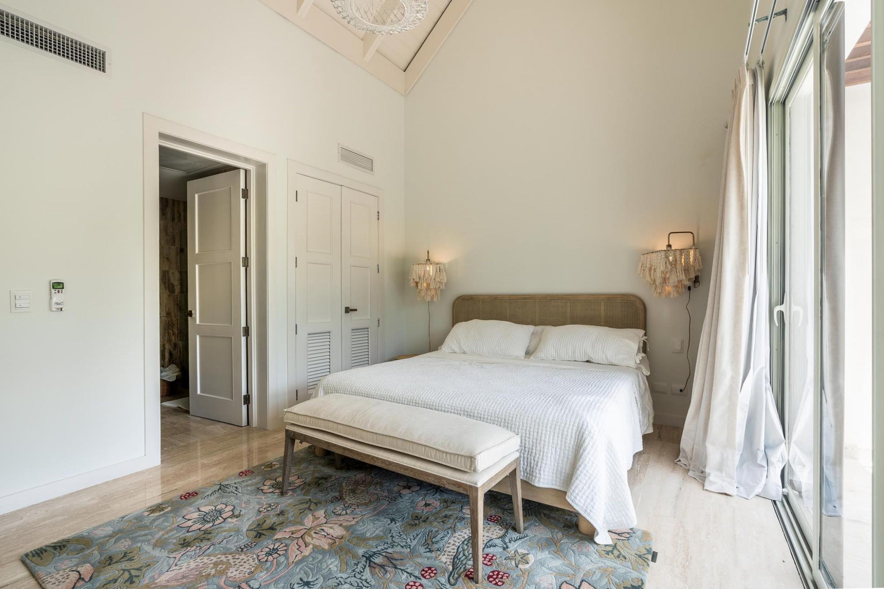 Une chambre à coucher avec un grand lit, un banc, un tapis à motifs et une vue sur un jardin par la fenêtre.