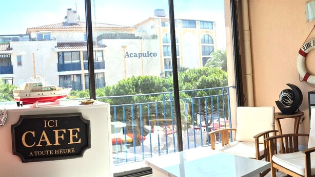 Een balkon met een tafel en stoelen, een bordje met de tekst 'ICI CAFÉ A TOUTE HEURE' en een uitzicht op gebouwen, waaronder Acapulco.