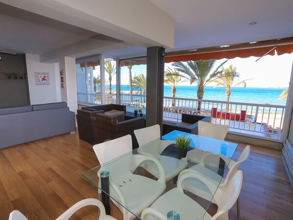 Un vaste espace de vie comprenant une table et des chaises en verre, un canapé et un balcon offrant une vue sur la plage et l’océan.