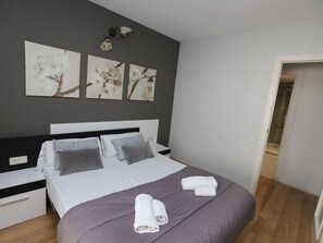 Un lit bien fait, avec une couvre-lit violette et des oreillers blancs, une table de chevet avec une lampe, et trois œuvres d’art florales accrochées au mur au-dessus du lit.