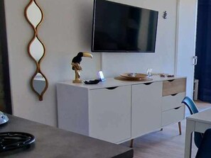 A modern hotel room with a flat-screen TV mounted on the wall, a wooden cabinet with a decorative mirror, a desk with a chair, and a small table with a lamp. - Charming 2-Room Apartment with Pool, Terrace, and A/C - 4 Sleeps - Near Port and Beach (Port Camargue)
