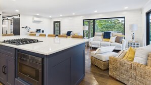 Et moderne køkken med en stor ø, et spiseområde med en hvid sofa og udsigt til grønne omgivelser.