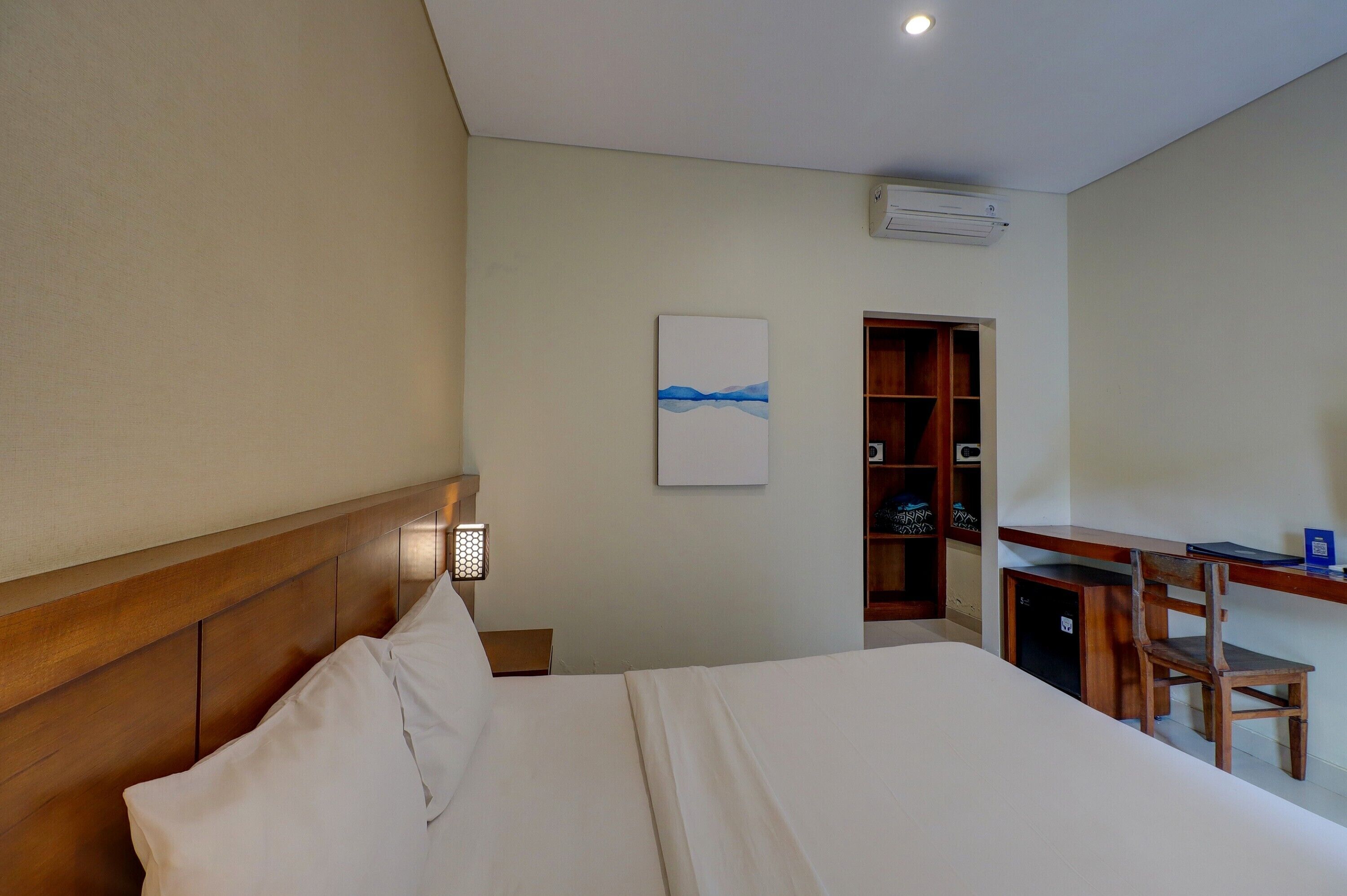 A hotel room with a wooden headboard, a bed with white linens, a desk with a chair, a small cabinet, and a wall-mounted air conditioner.