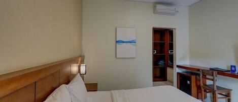 A hotel room with a wooden headboard, a bed with white linens, a desk with a chair, a small cabinet, and a wall-mounted air conditioner.