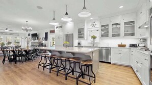 A modern kitchen with a central island, bar stools, white cabinetry, and a large window overlooking the city. - Resort Pool Home | Walk to Beach | 7 BR | Dogs (South Haven)