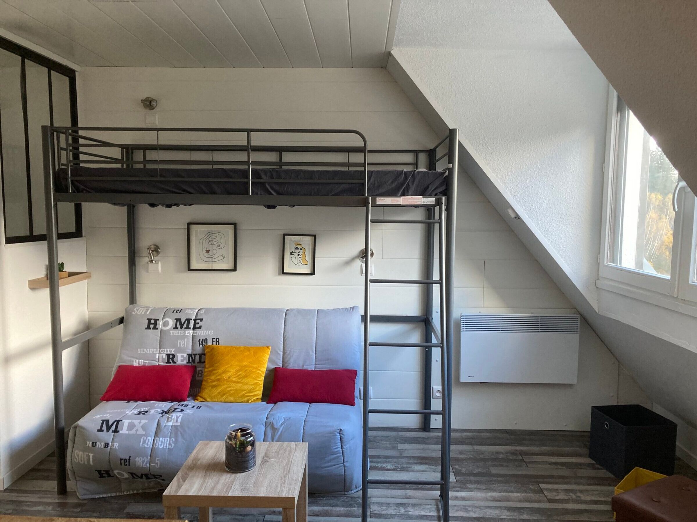A loft bedroom with a bunk bed, a sofa with colorful cushions, a small table, and a ladder.