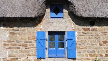Un bâtiment en pierre avec un toit de chaume et une petite fenêtre ornée de volets bleus.