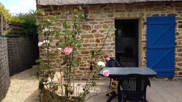 Une maison de campagne en pierre, avec un toit de chaume, un volet bleu et une table entourée de chaises à l’extérieur.