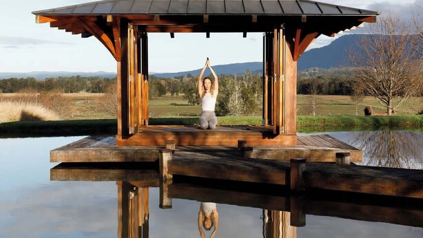 Een houten gazebo met een metalen dak dat uitkijkt over een wateroppervlak, met een persoon die een yoga-houding uitvoert in het midden.