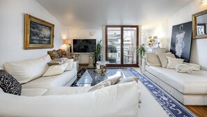 A living room with white sofas, a coffee table, and a TV. There is a painting on the wall and a window with a view of buildings. - Luxus penthouse in great location (Helsinki)