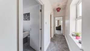 A hallway with a white door leading to a bathroom, a window with a plant, and a ceiling light fixture. - Spacious One bed, Stratford, Westfields, London (London)
