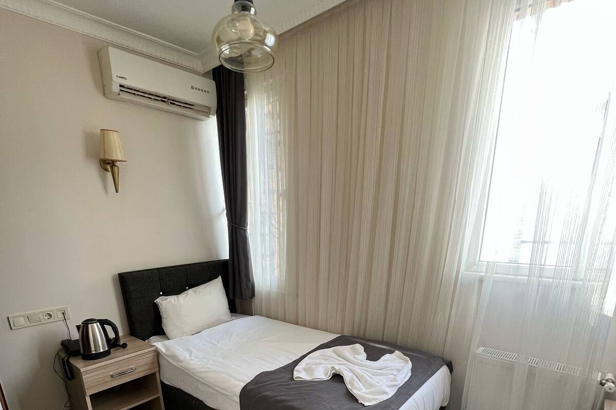 Quarto de hotel com uma cama, uma mesa de cabeceira com chaleira, uma cadeira e uma janela com cortinas leves.