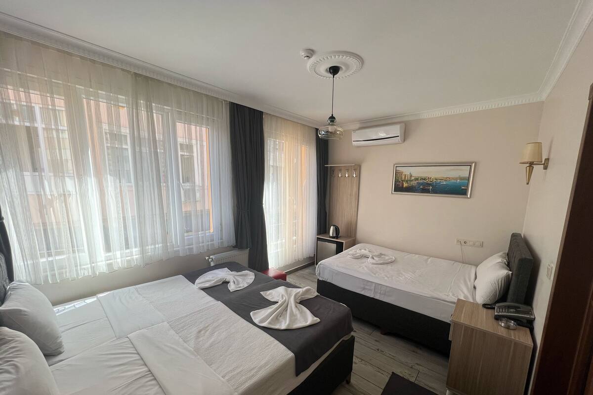 Quarto de hotel com duas camas, um ventilador de teto, ar condicionado, um quadro na parede e uma pequena mesa com chaleira.
