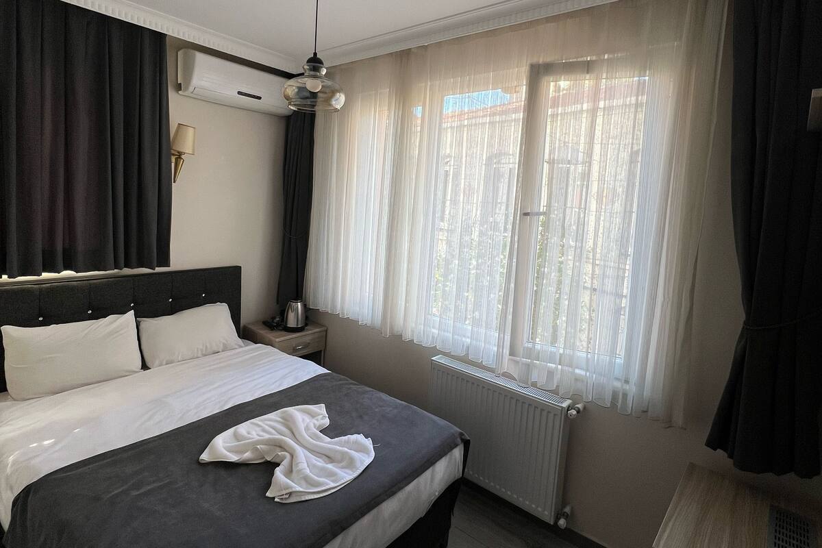 Quarto de hotel com cama, mesa de cabeceira, ar condicionado, janela com cortinas finas e piso de madeira.
