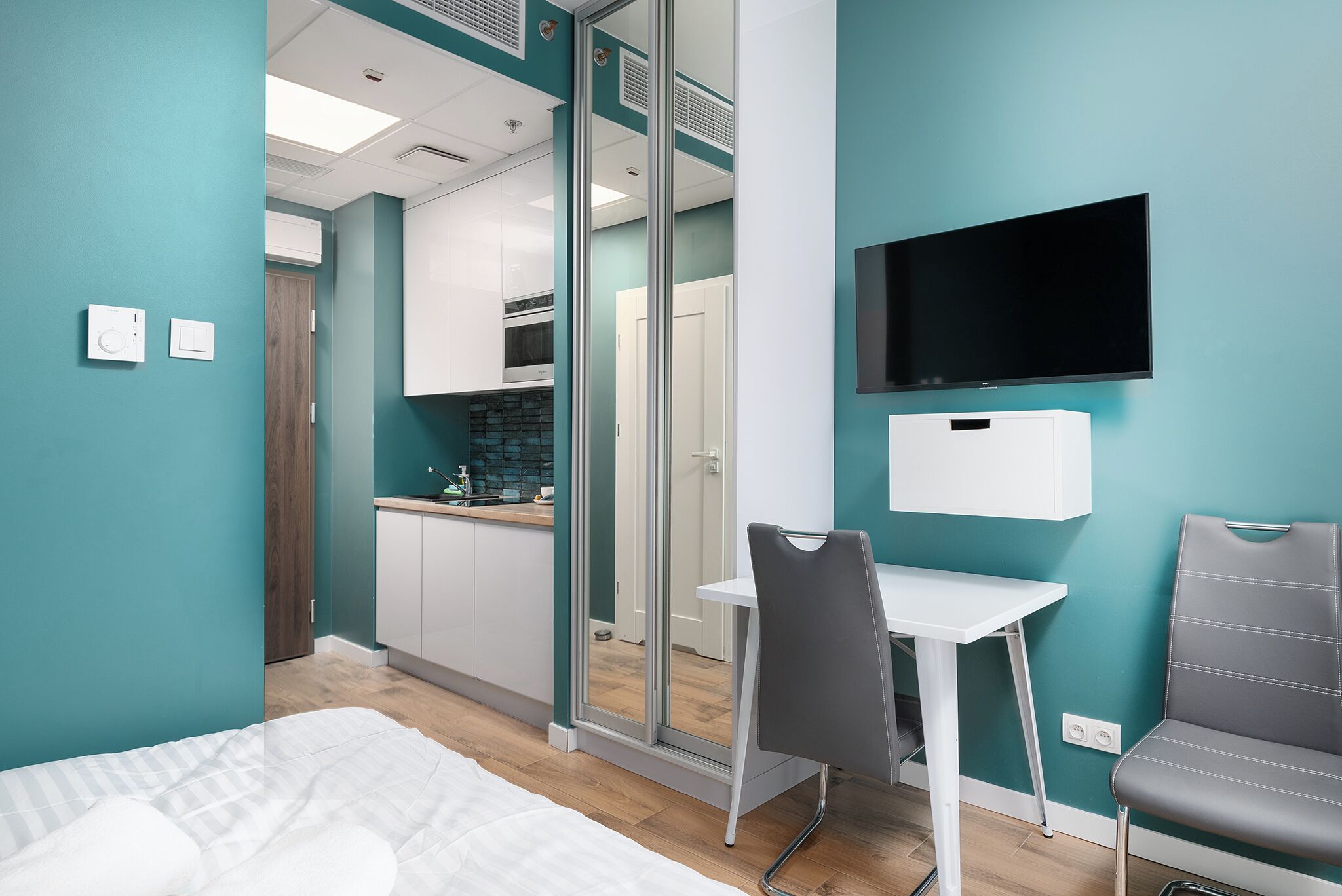 A modern hotel room with teal walls, a bed, a desk, a chair, a TV, and a kitchenette in the background.