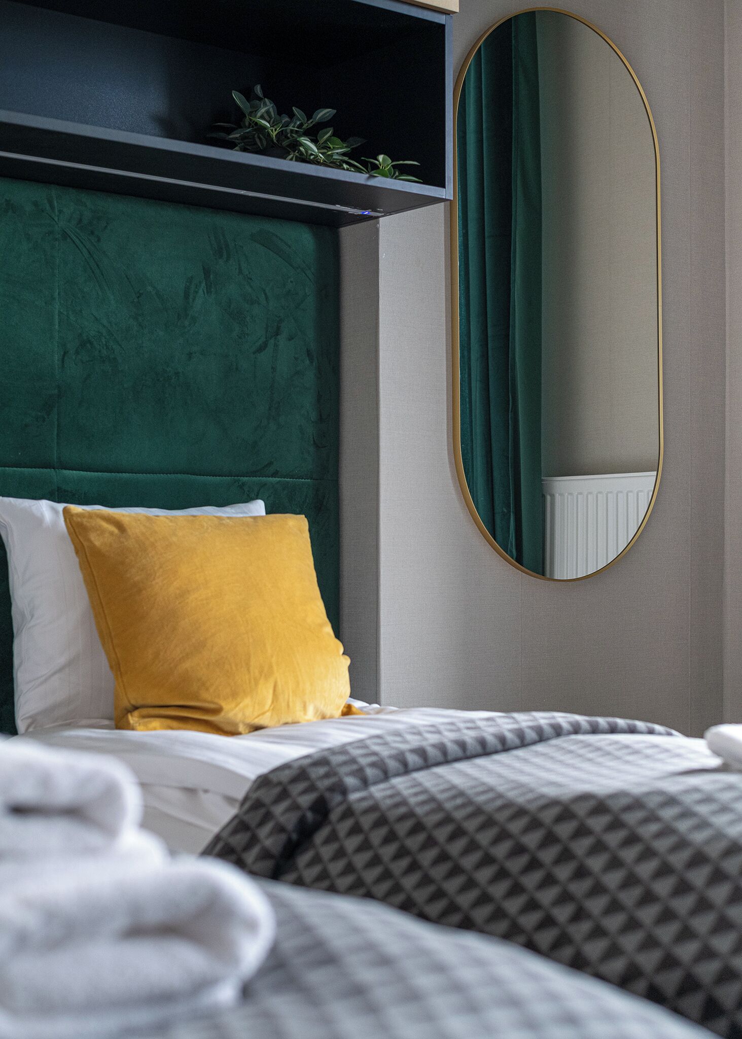 A neatly made bed with a green upholstered headboard, a yellow pillow, and a white pillow. A round mirror with a gold frame hangs on the wall.