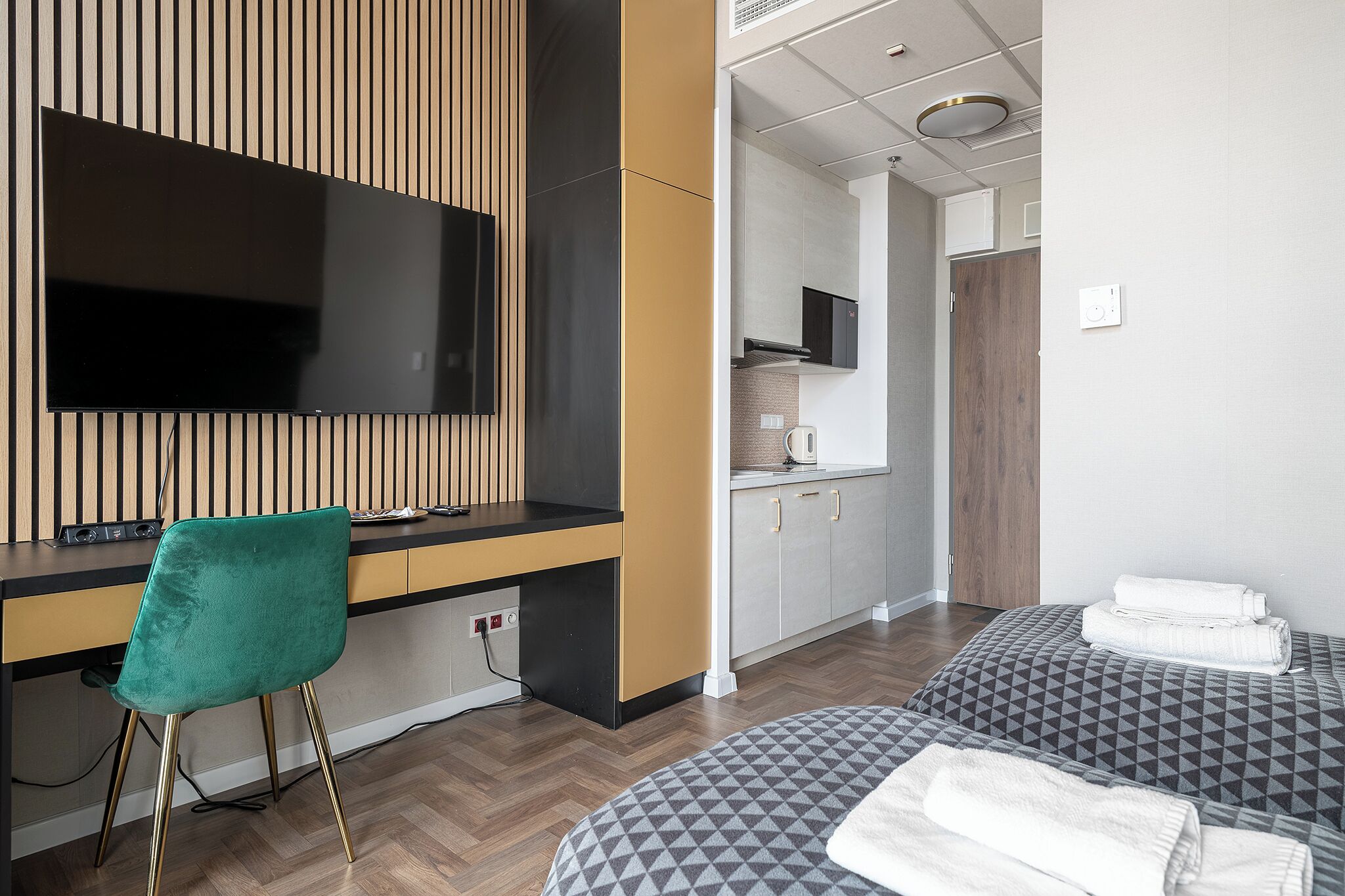 Una habitación de hotel moderna con televisión de pantalla plana, un escritorio con silla, una kitchenette y dos camas con toallas.