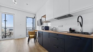 A modern kitchen with a large window, a dining area with a chair, and a city view. - RentPlanet - Studio Kasprzaka (Warsaw)